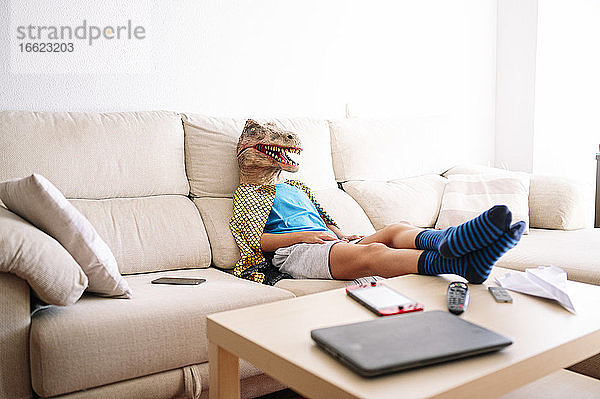 Junge mit Dinosauriermaske entspannt sich zu Hause auf dem Sofa