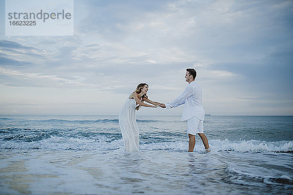 Verspieltes Paar hält sich an den Händen und steht im Wasser am Strand