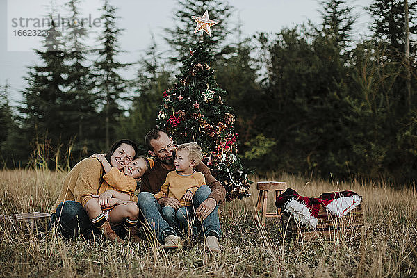 Lächelnde Familie sitzt am Weihnachtsbaum auf einer Wiese auf dem Lande bei Sonnenuntergang