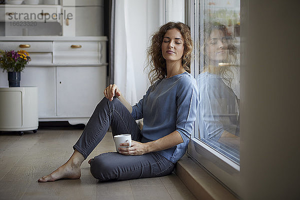 Mittlere erwachsene Frau mit Kaffeetasse  die sich zu Hause an eine Glastür lehnt