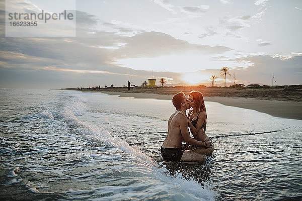 Paar in Badekleidung küsst sich bei Sonnenuntergang am Strand sitzend