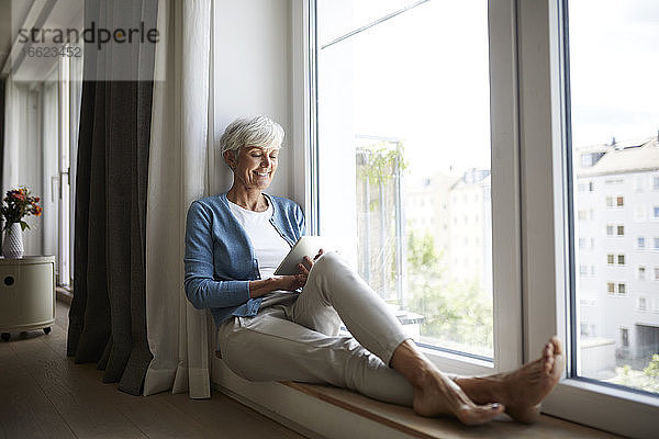 Glückliche ältere Frau  die ein digitales Tablet benutzt  während sie zu Hause am Fenster sitzt