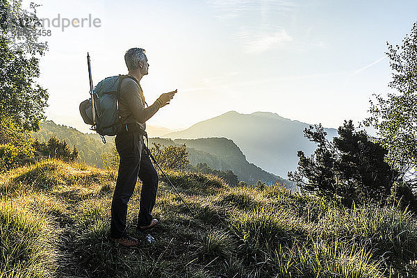 Mann  der ein Mobiltelefon benutzt  während er auf einem Berg gegen den Himmel bei Sonnenaufgang steht  Orobie  Lecco  Italien