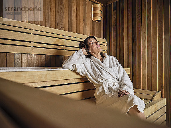 Ältere Frau mit Händen hinter dem Kopf entspannt sich in der Sauna im Wellnessbereich