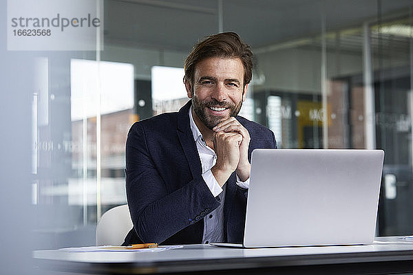 Lächelnder Geschäftsmann  der einen Laptop benutzt und auf einem Stuhl im Büro sitzt