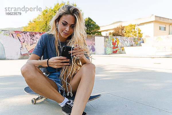 Junge blonde Frau  die ein Smartphone benutzt  während sie mit einem Skateboard im Park sitzt