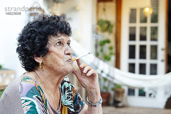 Nahaufnahme einer älteren Frau  die vor einem Haus im Hof sitzt und eine Zigarette raucht