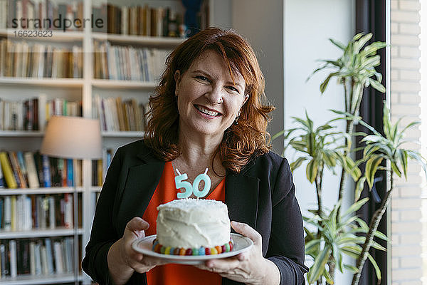 Frau hält Kuchen mit Kerze Nummer 50  während sie zu Hause steht
