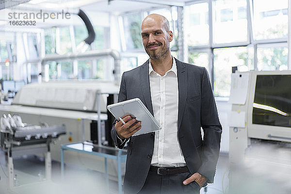 Lächelnder  selbstbewusster Geschäftsmann  der ein digitales Tablet hält  während er mit der Hand in der Tasche in einer hellen Fabrik steht
