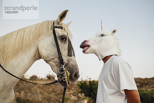 Weißes Pferd und Mann mit Einhornmaske schauen sich an