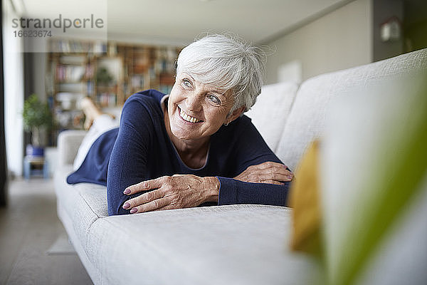 Lächelnde ältere Frau  die mit verschränkten Armen wegschaut  während sie zu Hause auf dem Sofa liegt