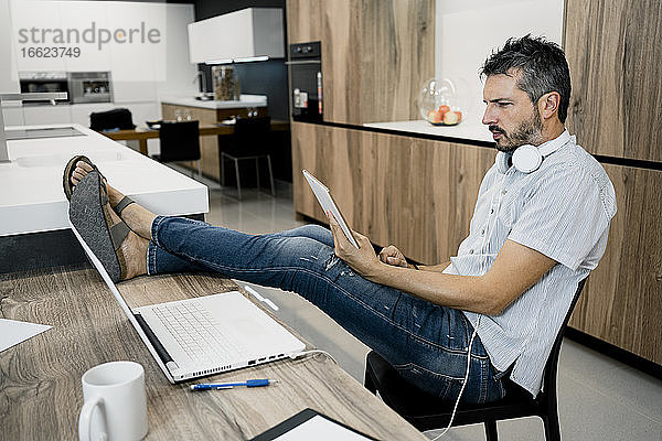 Älterer männlicher Freiberufler  der ein digitales Tablet hält  während er mit hochgelegten Füßen auf einem Schreibtisch im Büro sitzt