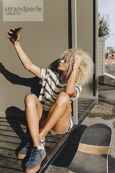 Blonde Frau  die ein Selfie mit ihrem Smartphone macht  während sie an einer Metallwand sitzt