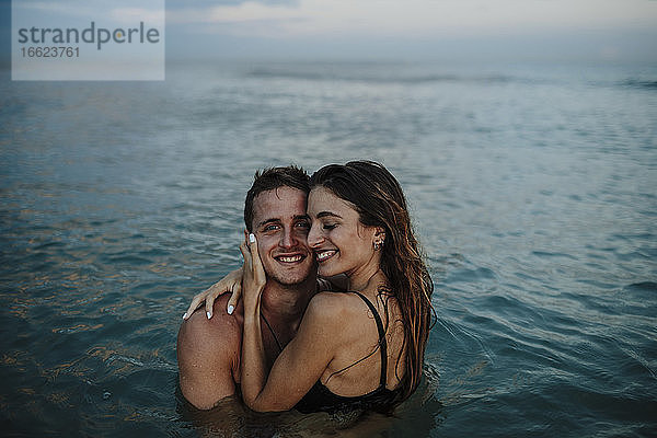 Lächelndes Paar  das sich umarmt  während es bei Sonnenuntergang am Strand im Wasser steht