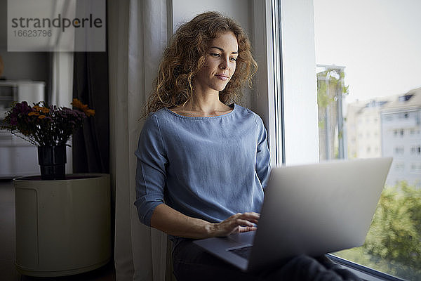 Frau benutzt Laptop  während sie zu Hause auf der Fensterbank sitzt