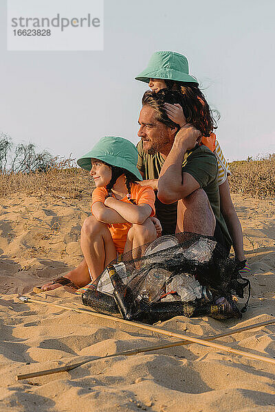 Vater und Töchter sitzen auf Sand mit Müllsack