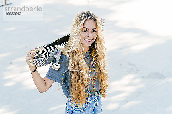 Lächelnde blonde Frau  die ein Skateboard hält  während sie im Park steht