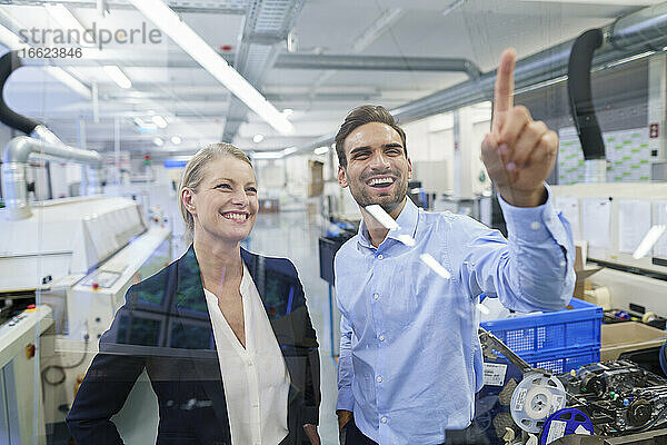 Glücklicher junger männlicher Techniker  der auf eine grafische Benutzeroberfläche auf einem Glas in einer Fabrik zeigt