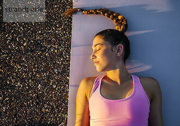 Junge Frau mit geschlossenen Augen meditiert im Liegen auf einer Matte bei Sonnenuntergang