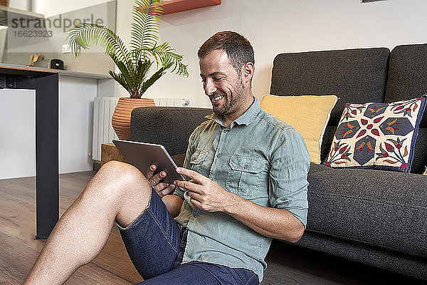 Lächelnder Mann  der ein digitales Tablet benutzt  während er zu Hause auf dem Sofa sitzt