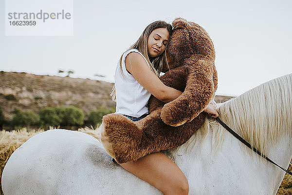 Porträt einer schönen jungen Frau  die einen großen Teddybären umarmt  während sie mit geschlossenen Augen auf einem Pferd sitzt
