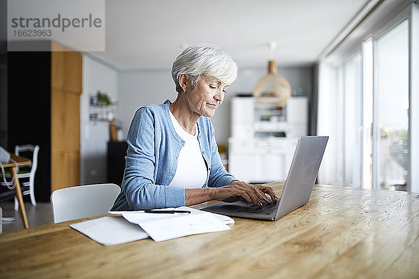 Ältere Frau arbeitet am Laptop  während sie zu Hause auf einem Stuhl sitzt