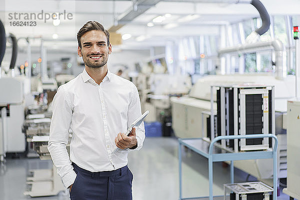 Lächelnder  selbstbewusster junger Geschäftsmann  der ein digitales Tablet hält  während er mit den Händen in den Taschen in einer beleuchteten Fabrik steht