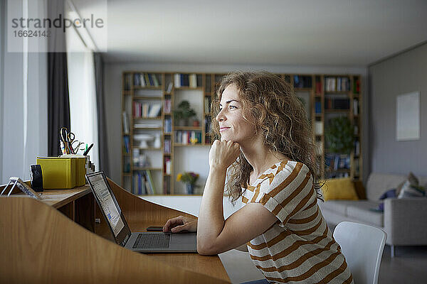 Lächelnde Frau mit dem Kopf in den Händen sitzt am Schreibtisch zu Hause