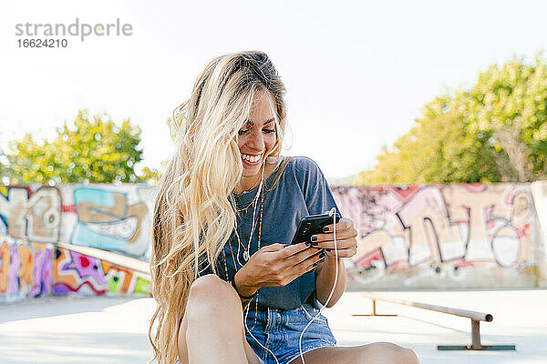 Glückliche junge blonde Frau  die ihr Smartphone benutzt  während sie mit einem Skateboard im Park sitzt