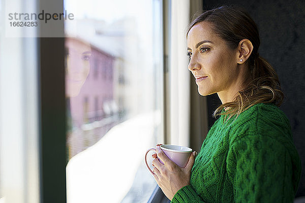Frau hält Kaffeetasse  während sie zu Hause am Fenster steht