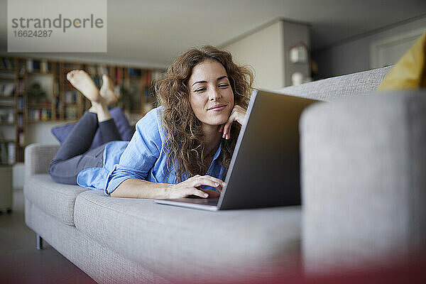 Lächelnde Frau  die einen Laptop benutzt  während sie zu Hause auf dem Sofa liegt