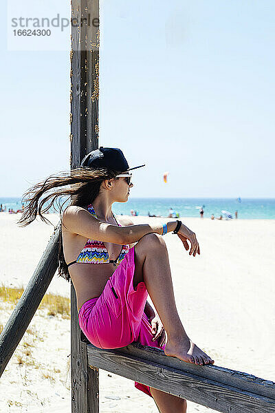 Junge Frau im Bikini auf einem Geländer am Strand sitzend gegen den klaren Himmel