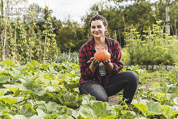 Lächelnde junge Frau hält Kürbis  während sie inmitten von Pflanzen im Gemüsegarten kniet