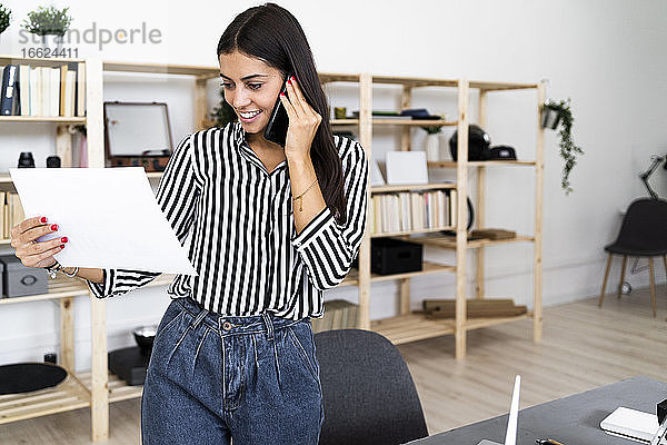 Lächelnde junge Architektin  die mit ihrem Smartphone spricht  während sie einen Plan im Büro betrachtet
