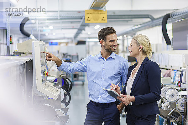 Lächelnder männlicher Techniker  der eine Geschäftsfrau ansieht  während er in einer beleuchteten Fabrik vor Maschinen steht