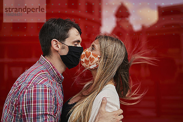Küssendes Paar mit Gesichtsschutzmaske an einer Acrylwand