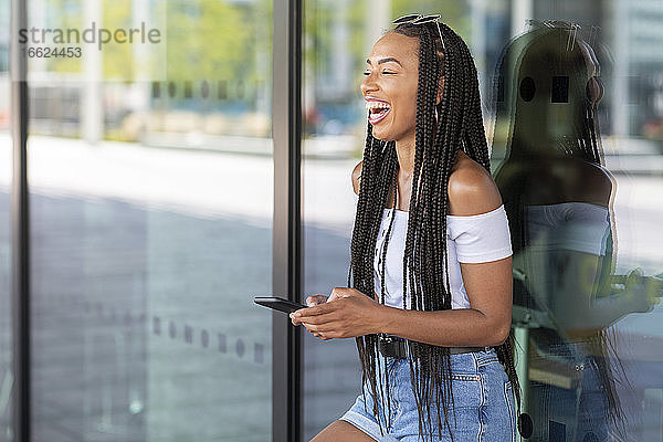 Frau lacht  während sie ein Mobiltelefon gegen eine Glaswand in der Stadt benutzt