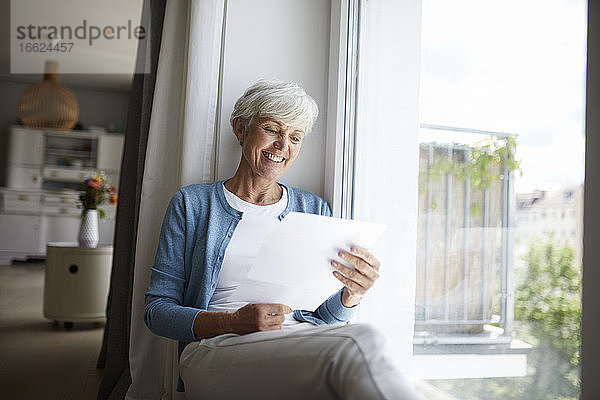 Ältere Frau liest fröhlich einen Brief  während sie zu Hause am Fenster sitzt