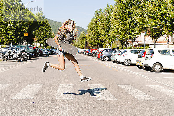 Fröhliche junge Frau  die ein Skateboard hält und über einen Zebrastreifen in der Stadt springt