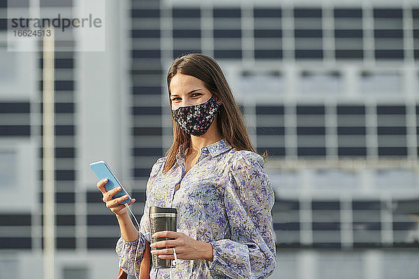 Junge Frau mit Gesichtsmaske  die eine Thermoskanne hält  während sie ein Mobiltelefon in der Stadt benutzt