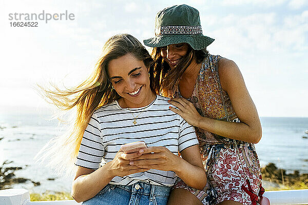 Lächelnde junge Freundinnen benutzen ihr Smartphone am Meer während eines sonnigen Tages