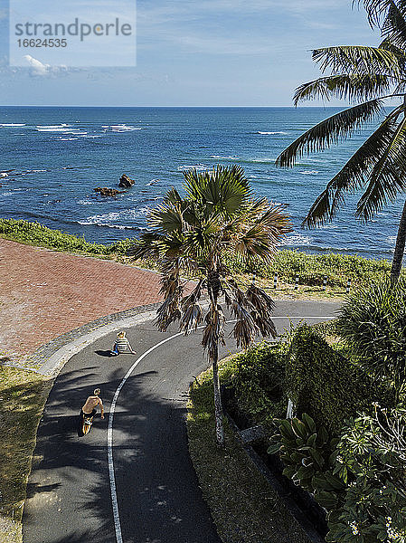 Indonesien  Bali  Zwei junge Männer auf dem Skateboard entlang der Küstenstraße mit dem Meer im Hintergrund
