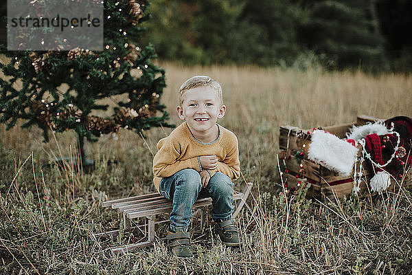 Niedlicher Junge sitzt auf Schlitten gegen Weihnachtsbaum auf dem Lande