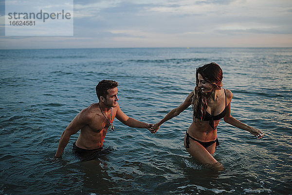 Paar hält sich an der Hand  während es bei Sonnenuntergang am Strand im Wasser spazieren geht