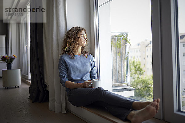Frau hält Kaffeetasse  während sie auf der Fensterbank zu Hause sitzt
