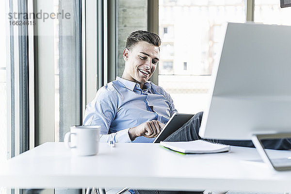 Lächelnder Geschäftsmann mit digitalem Tablet am Schreibtisch im Büro