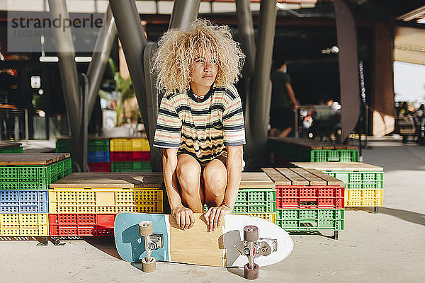 Afroblonde Frau hält Skateboard und schaut weg  während sie auf einem Kistensitz an einem sonnigen Tag sitzt