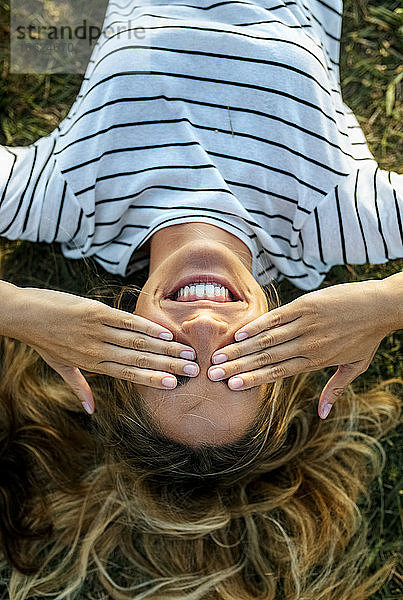 Glückliche junge Frau  die ihre Augen mit den Händen bedeckt  während sie sich auf einem Feld entspannt