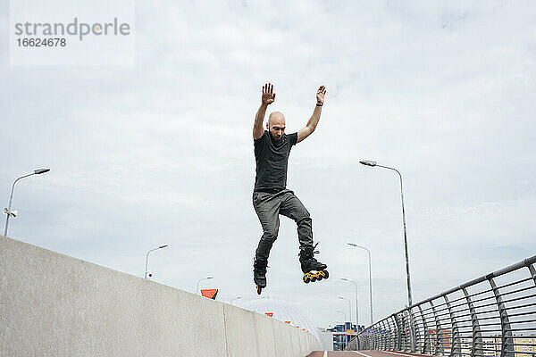 Junger Mann mit Inline-Skates springt auf Brücke gegen Himmel