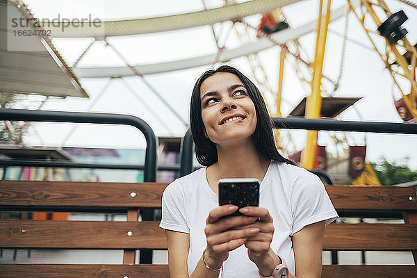 Junge Frau benutzt ihr Smartphone  während sie auf einer Bank im Vergnügungspark sitzt